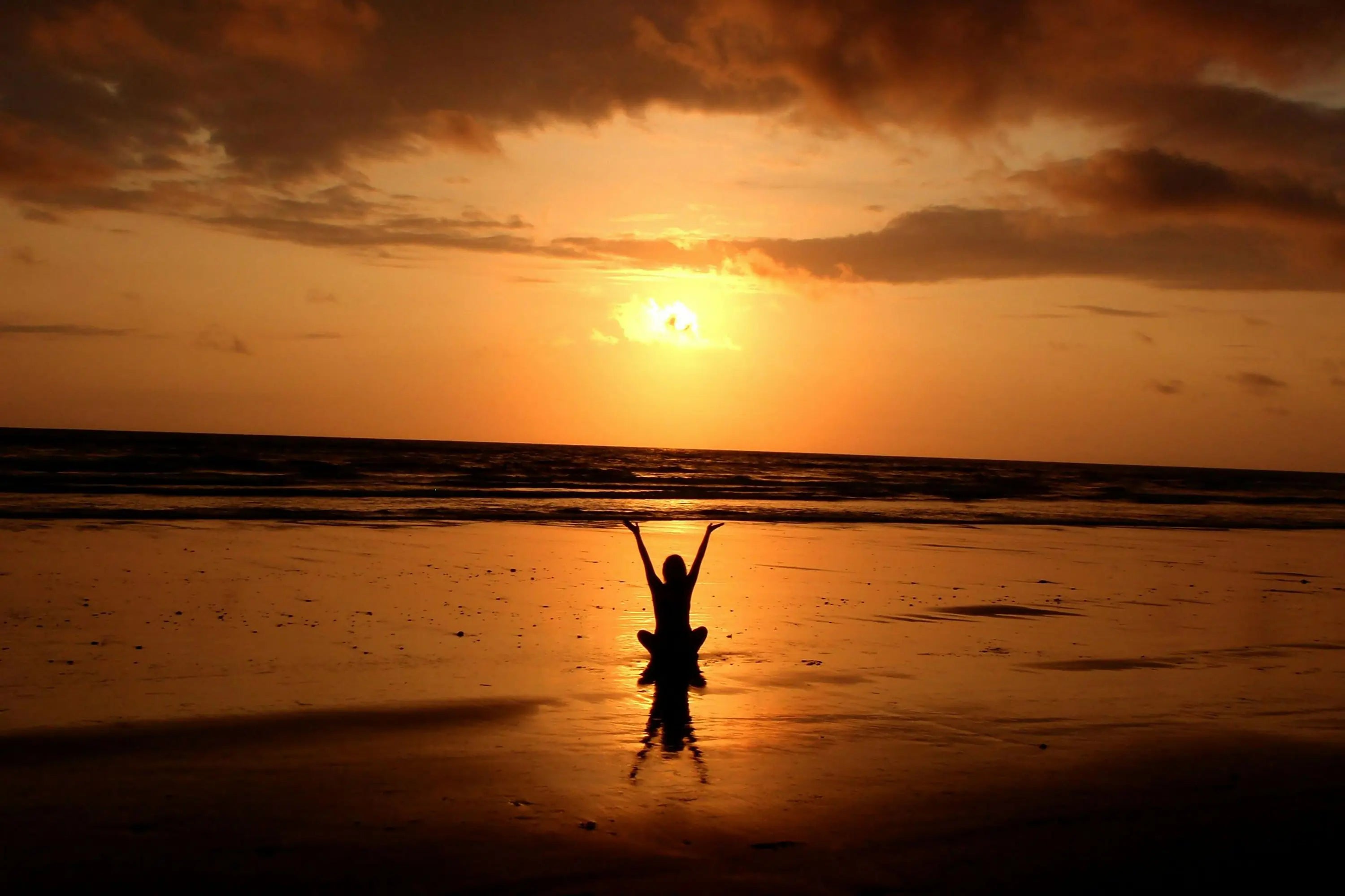 a silhouette of a person sitting on the beach and watching the sunset with his hands up